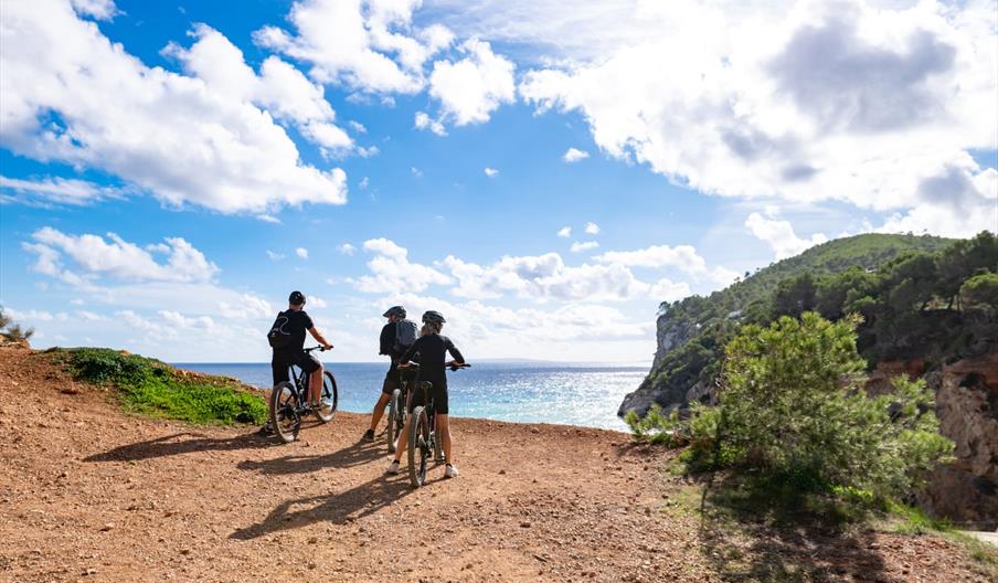 Personas recorriendo una ruta en bicicleta con vistas a la costa y los acantilados, en un paisaje natural de Ibiza bajo un cielo luminoso.
