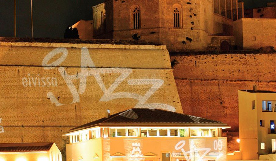 La fachada de la muralla de Dalt Vila refleja el emblema de Eivissa Jazz en color blanco. Al fondo se ve la Catedral de Santa María y, a lo spies, par