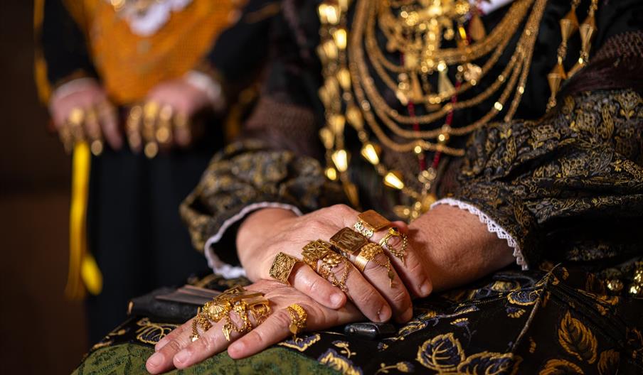 Una mujer lleva muchos anillos dorados y grandes en las manos y muchos collares en el cuello. Son las E
emprendades.