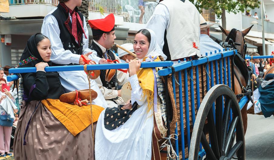 Tres hombres y dos mujeres, una de ellas es una niña, viajan en un carro azul de grandes ruedas de madera tirado por caballo. Todos van ataviados con