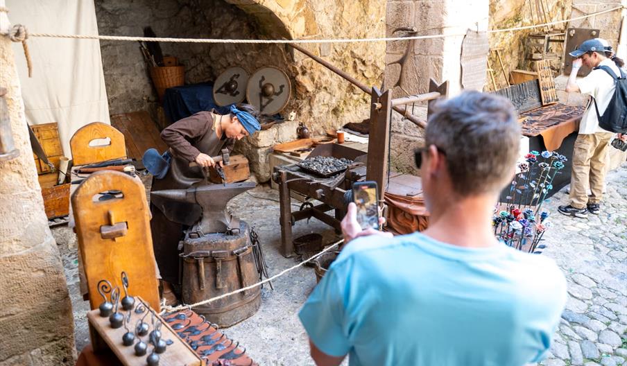 Persona ataviada con ropas medievales trabaja el acero durante Fira Medieval en las calles de Dalt Vila, mientras un visitante fotografía o graba la e