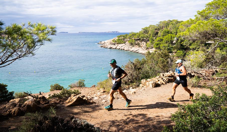 Dos atletas corren por un camino en la naturaleza en la Ibiza Trail Marathon Festival, con el mar Mediterráneo al fondo.