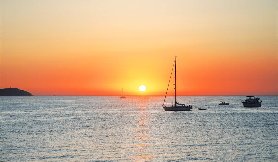 Puesta de sol en Ibiza con un velero y varios barcos más en el Mediterráneo. Colores anaranjados en el cielo mientras el sol se oculta.