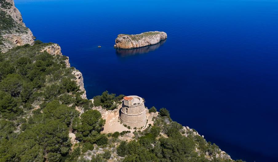 Vista aérea de la Torre des Molar sobre un acantilado del norte de Ibiza, rodeada de pinares y con el mar Mediterráneo y un islote rocoso al fondo.