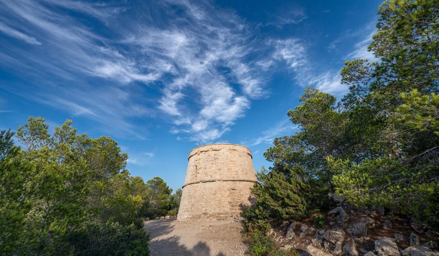 Torre de Portinatx, antigua torre de defensa costera de piedra rodeada de bosque mediterráneo bajo un cielo azul con nubes en el norte de Ibiza.