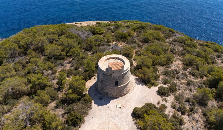 Vista aérea de la Torre d’en Valls, antigua torre de vigilancia costera rodeada de vegetación mediterránea y acantilados junto al mar en Ibiza.