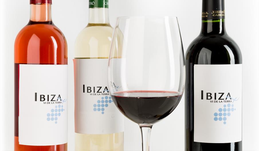 Botellas de vino tinto, blanco y rosado elaborados en bodegas de Ibiza, acompañadas por una copa servida, representando la tradición vitivinícola de l