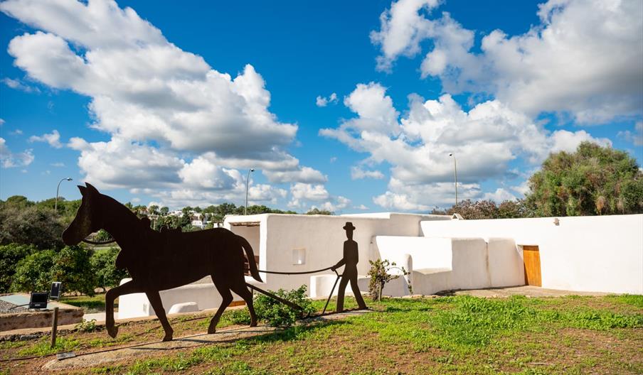 Estructura de metal con un hombre que anda cogido de las riendas de un caballo. Detrás se ve una construcción que sigue las líneas típicas de la arqui