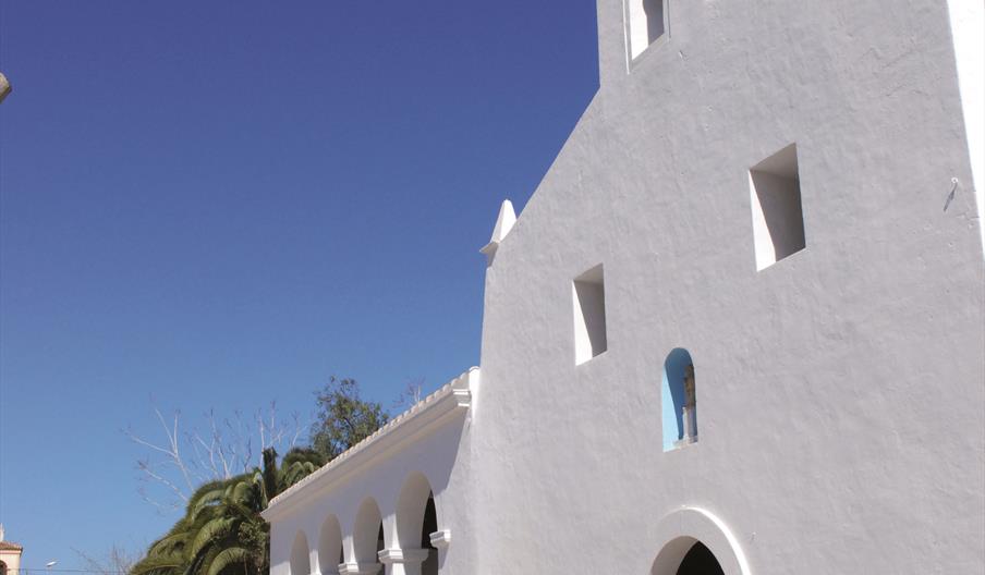 Iglesia de fachada blanca y arquitectura típica ibicenca con puerta pequeña y campanario pequeño arriba.Tiene dos ventanas pequeñas encima de la puert