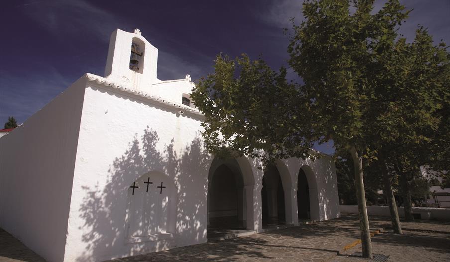 Se ve una iglesia totalmente blanca de estilo ibicenco, con tres cruces en la pared y un campanario. A la derecha aparecen tres arcos de medio punto q