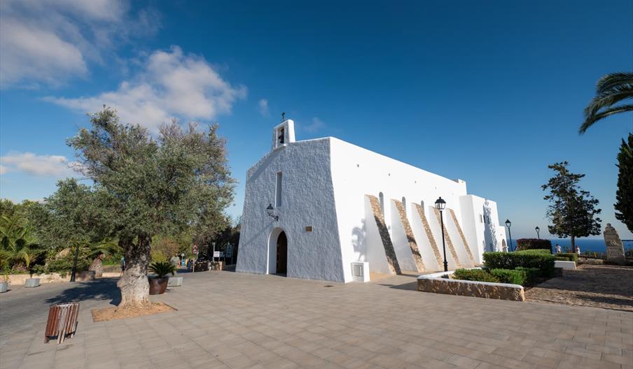 La iglesia de Es Cubells, un edificio blanco y austerio con contrafuertes blancos y un pequeño campanario enla fachada. La puerta es pequeña. Está sit