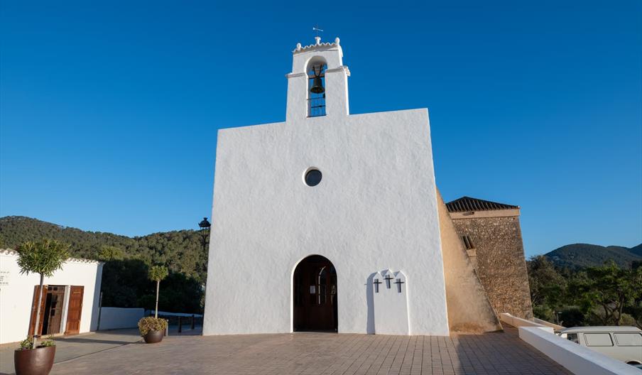 Fachada blanca y de estilo ibicenco con una puerta en forma de arco de medio punto pequeña. A la derecha sobre sale una placa blanca con tres cruces.