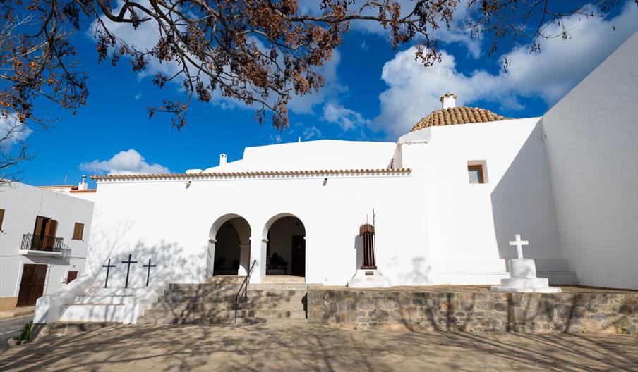 Amplia plaza que, a través de unos escalones, sube a una iglesia blanca típica ibicenca. A la izquierda aparecen tres cruces al final es unos escalone