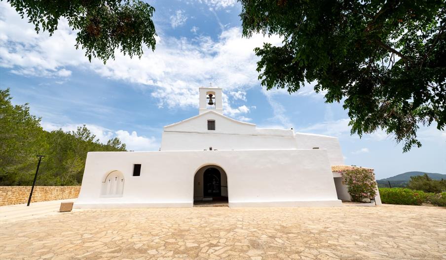 Iglesia tradicional ibicenca de paredes encaladas y arquitectura defensiva, rodeada de naturaleza bajo un cielo azul con nubes en Ibiza.