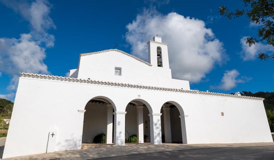Iglesia tradicional ibicenca de arquitectura blanca y porche con arcos, bajo un intenso cielo azul con nubes, en un entorno tranquilo de Ibiza.