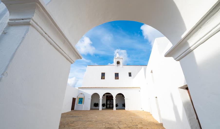 Patio interior de una iglesia tradicional ibicenca de paredes blancas y arcos, iluminado por el sol bajo un cielo azul con nubes en Ibiza.