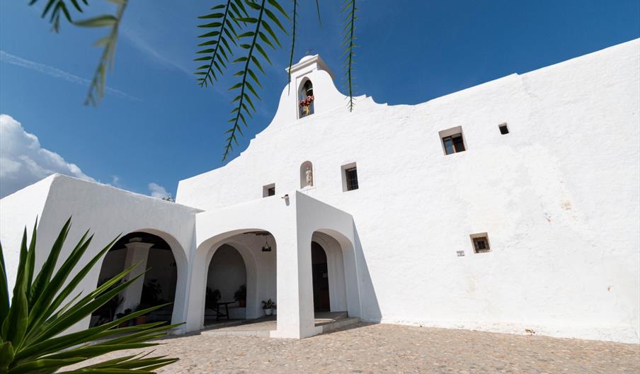 Fachada de la iglesia tradicional ibicenca de paredes encaladas y arcos, rodeada de vegetación mediterránea bajo un cielo azul despejado en Ibiza. Cor