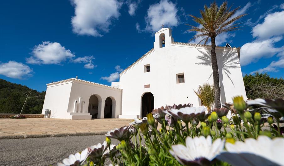 Iglesia de Sant Vicent de sa Cala, de arquitectura tradicional ibicenca y paredes blancas, rodeada de flores y palmeras bajo un intenso cielo azul en