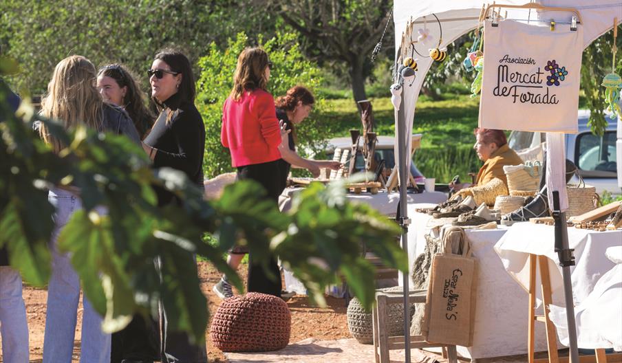 Personas visitando el Mercado de Forada entre puestos de artesanía y productos locales, en un entorno rural rodeado de naturaleza en el interior de Ib