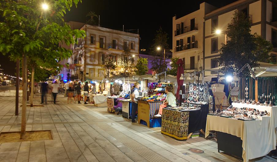 Puestos de artesanía, joyería y productos locales en el Mercadillo Hippy del Puerto de Ibiza, iluminado al anochecer en el paseo marítimo del casco ur