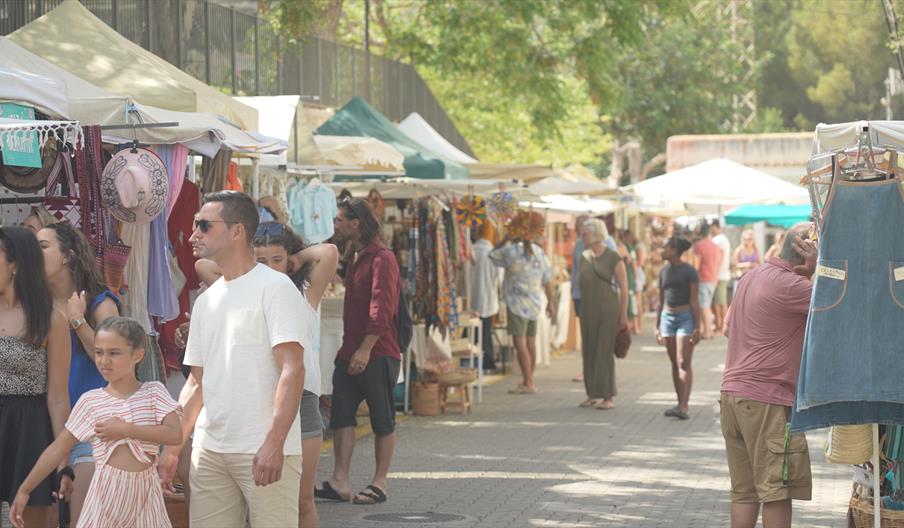 Visitantes recorriendo el mercadillo hippy de Ses Figueretes entre puestos de artesanía, moda y productos locales en un ambiente animado junto al mar