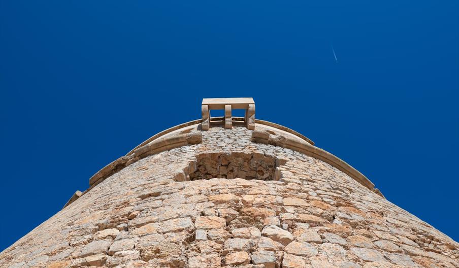 Detalle en contrapicado de la Torre de sa Sal Rossa, antigua torre de defensa costera construida en piedra bajo un intenso cielo azul en Ibiza.