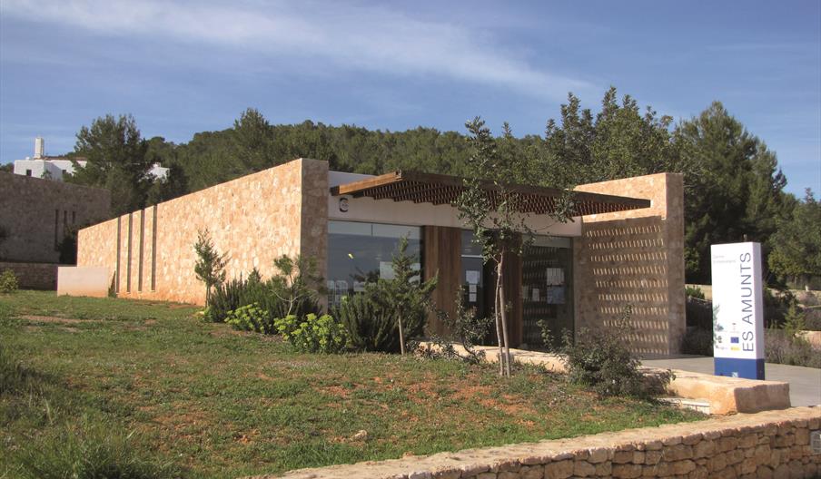 Edificio de piedra marrón claro con un jardín en l aentrada y rodeado de árboles. Hay un cartel blanco con las letras en azul donde reza "Es Amunts".
